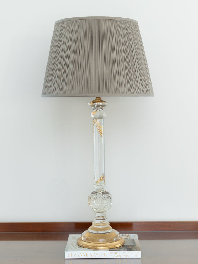 A classic table lamp with a pleated taupe-gray fabric shade, a clear glass column with gold accents, and a gold-finished base, resting on a book against a white wall.