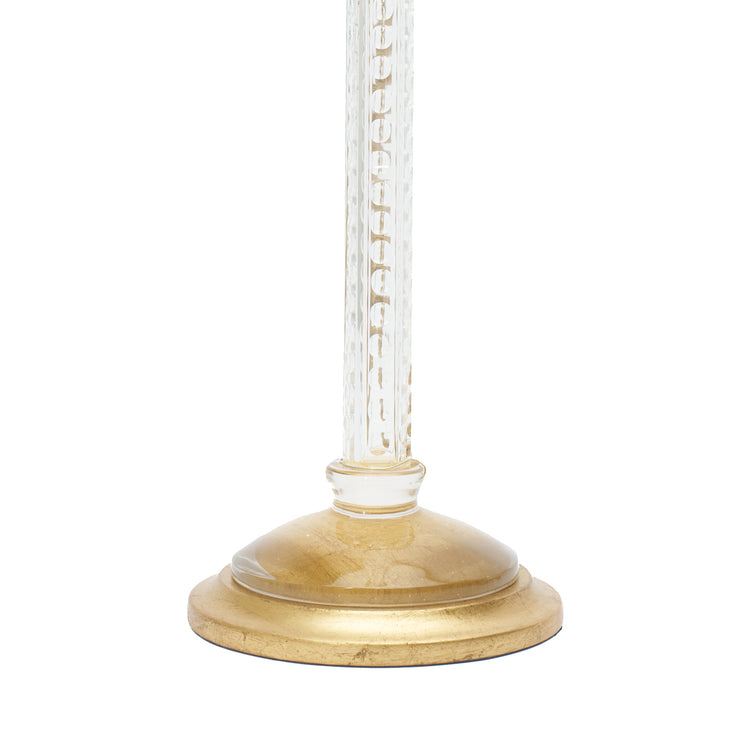 A close-up of a table lamp base, featuring a round gold-finished base and a clear glass stem, shown against a plain white background.