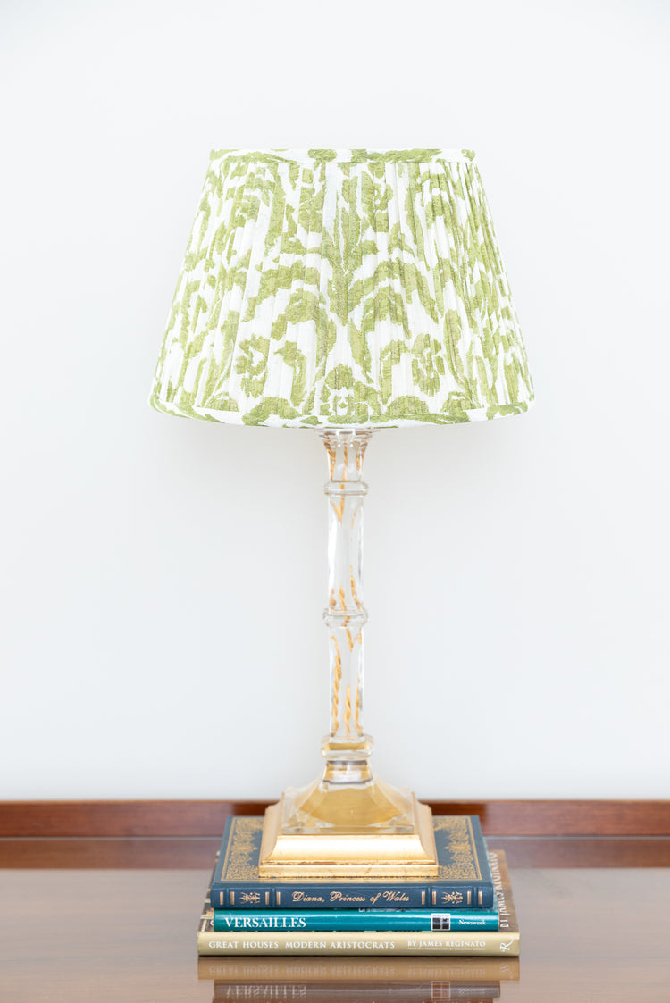 A table lamp with a pleated green and white shade, a clear glass stem, and a gold base, styled on top of two books against a white wall.
