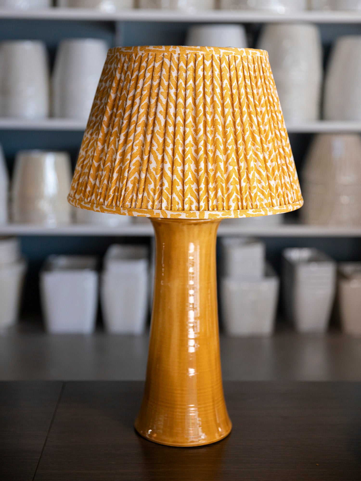A table lamp with a glossy mustard yellow ceramic base and a pleated fabric shade