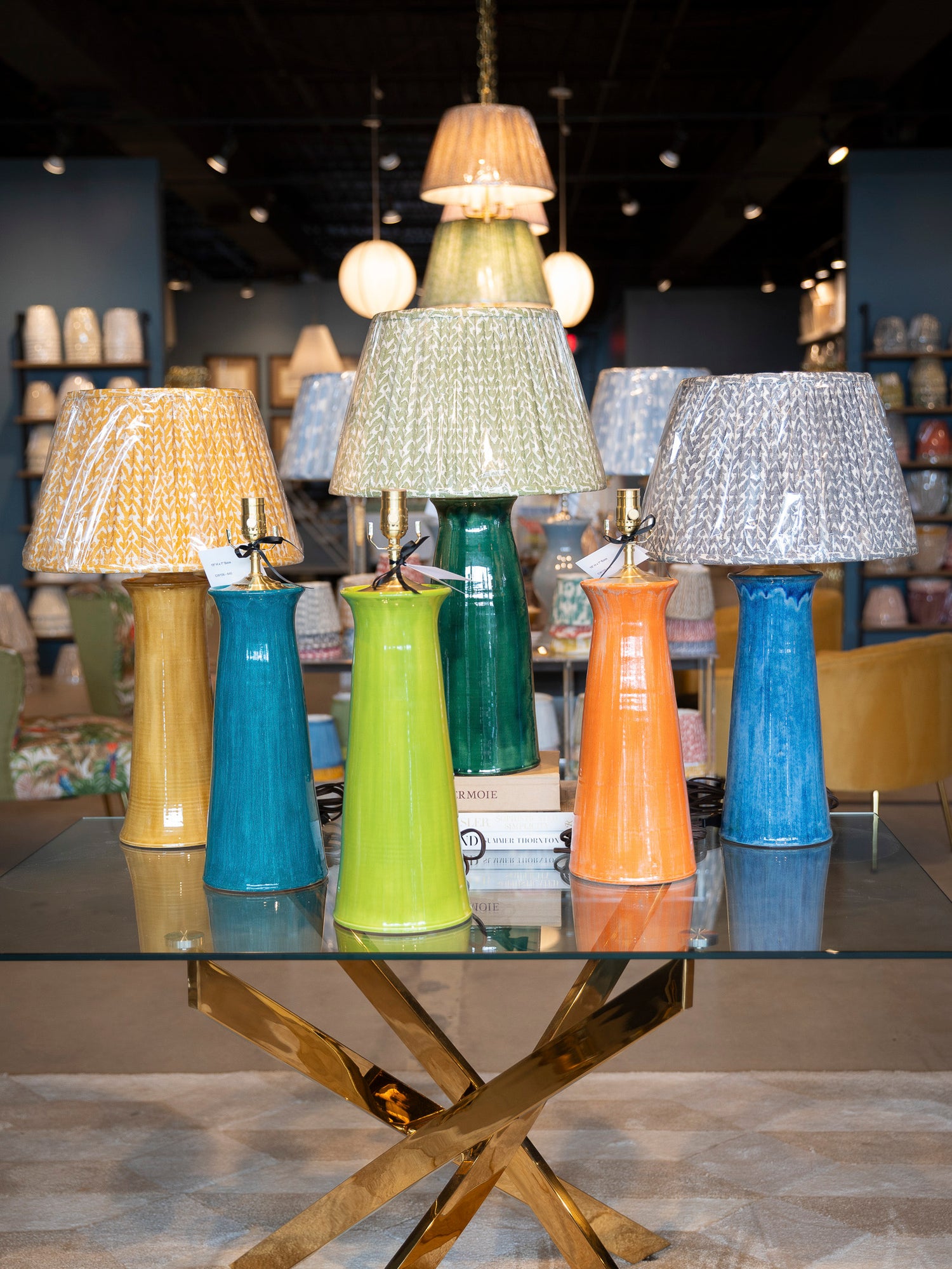 A collection of ceramic pottery table lamps with patterned fabric shades in yellow, blue, and green, displayed on a glass-topped table in a showroom.