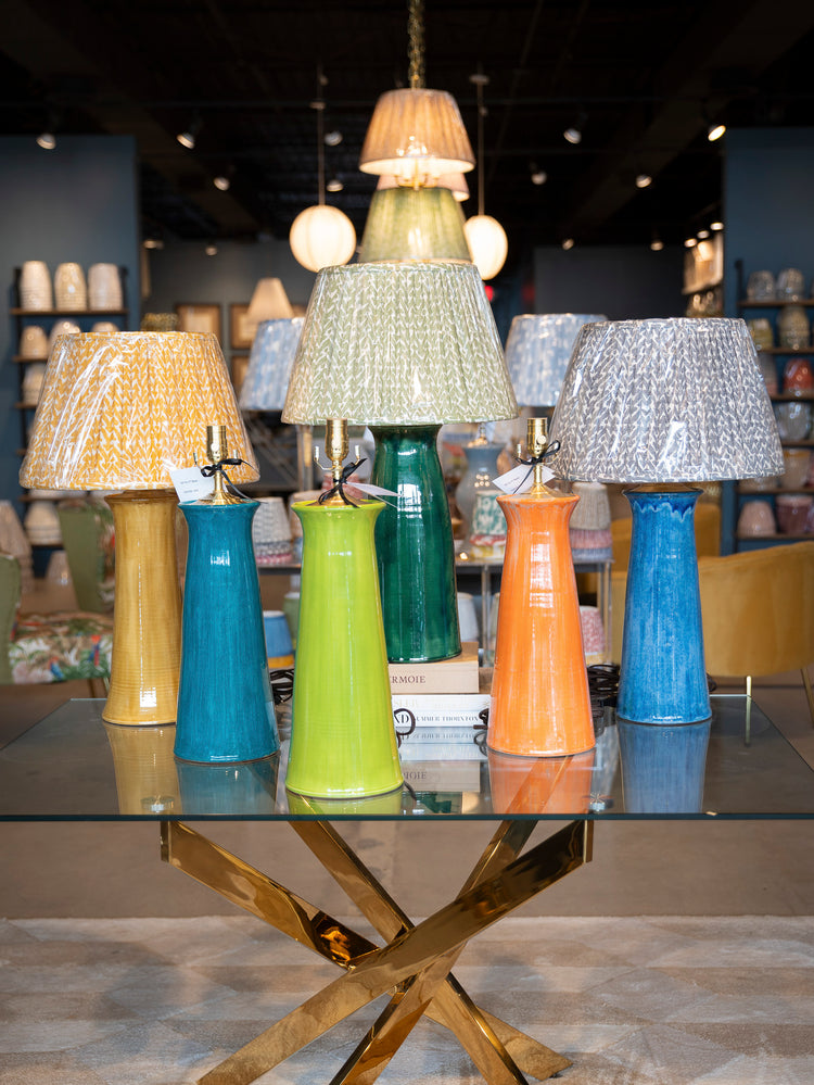 A collection of ceramic pottery table lamps with patterned fabric shades in yellow, blue, and green, displayed on a glass-topped table in a showroom.