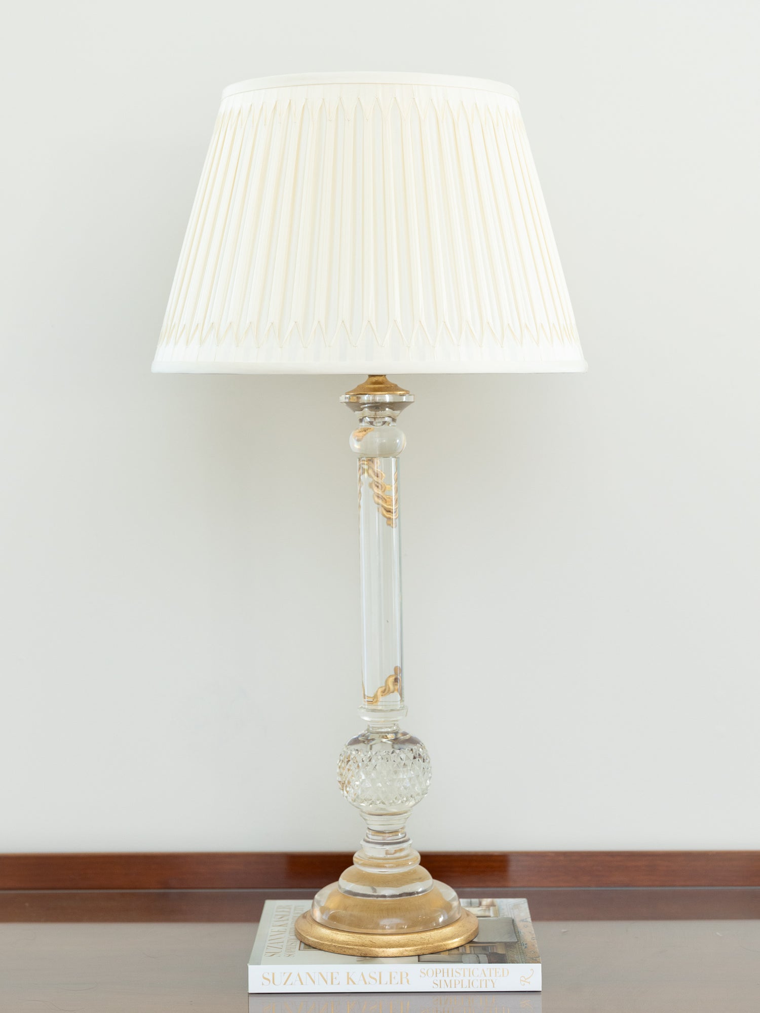 A classic table lamp with a pleated off-white fabric shade, a clear glass column with gold decorative accents, and a gold-toned base, resting on a stack of books against a neutral wall.