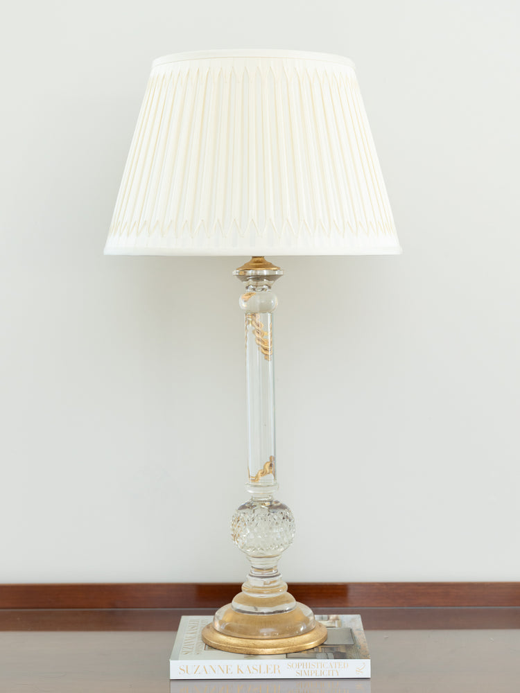 A classic table lamp with a pleated off-white fabric shade, a clear glass column with gold decorative accents, and a gold-toned base, resting on a stack of books against a neutral wall.