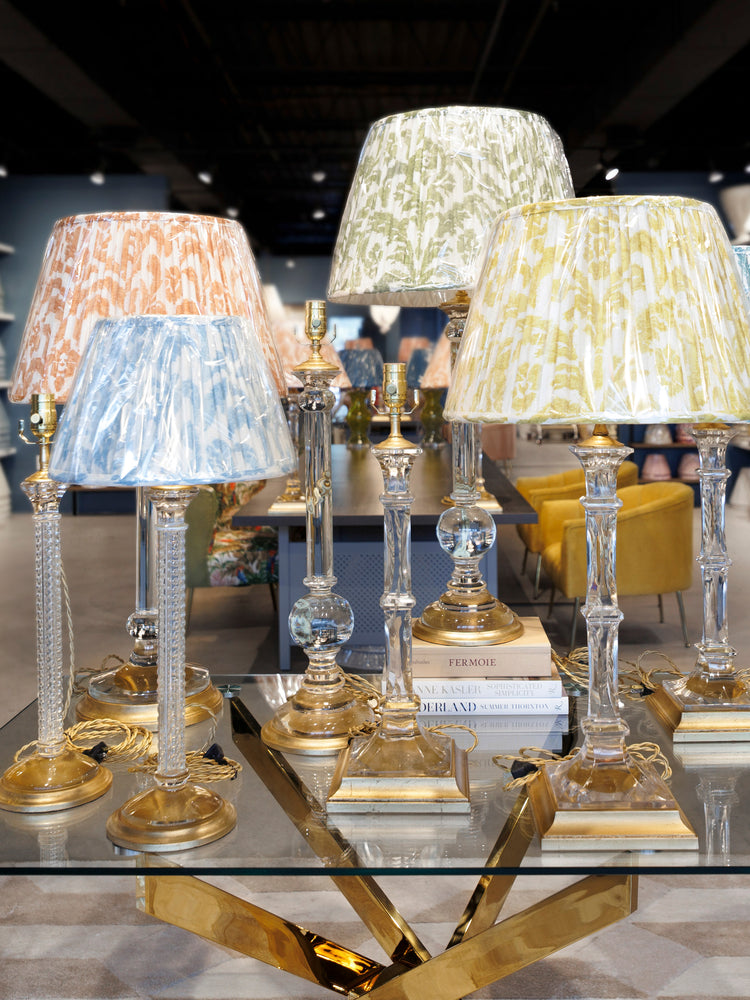 A collection of elegant table lamps with patterned fabric shades in blue, peach, and beige, paired with clear glass stems and ornate gold bases, displayed on a glass-topped table in a showroom.