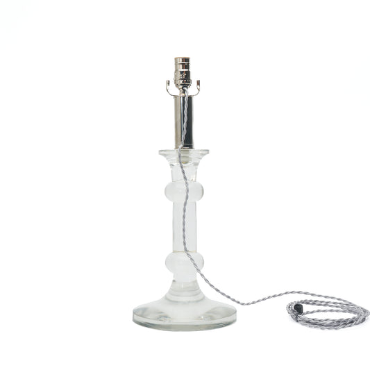 Clear glass table lamp base with a silver metal socket and a silver braided cord on a white background.