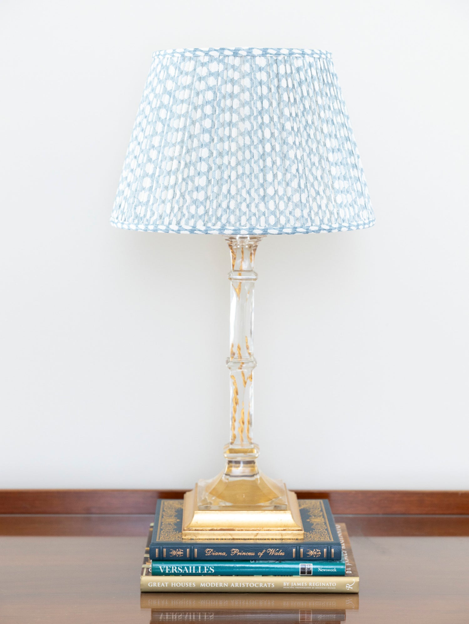 A table lamp with a pleated blue and white shade, a clear glass stem, and a gold base, styled on top of two books against a white wall.