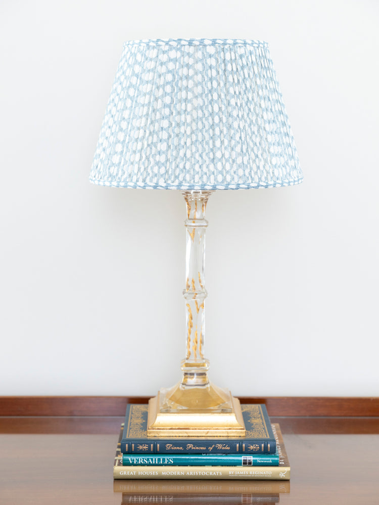 A table lamp with a pleated blue and white shade, a clear glass stem, and a gold base, styled on top of two books against a white wall.