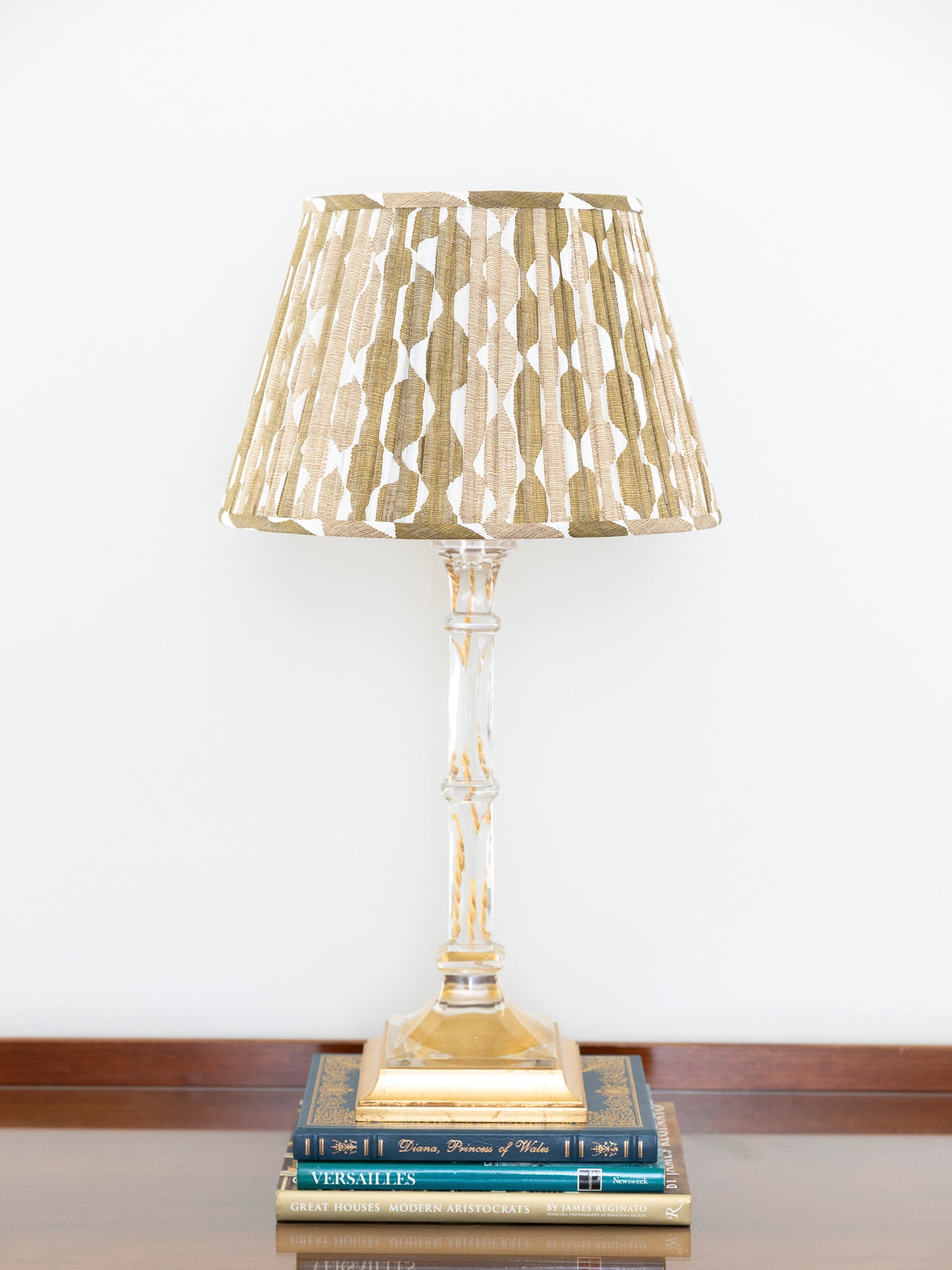 A table lamp with a pleated gold and beige shade, a clear glass stem, and a gold base, styled on top of two books against a white wall.