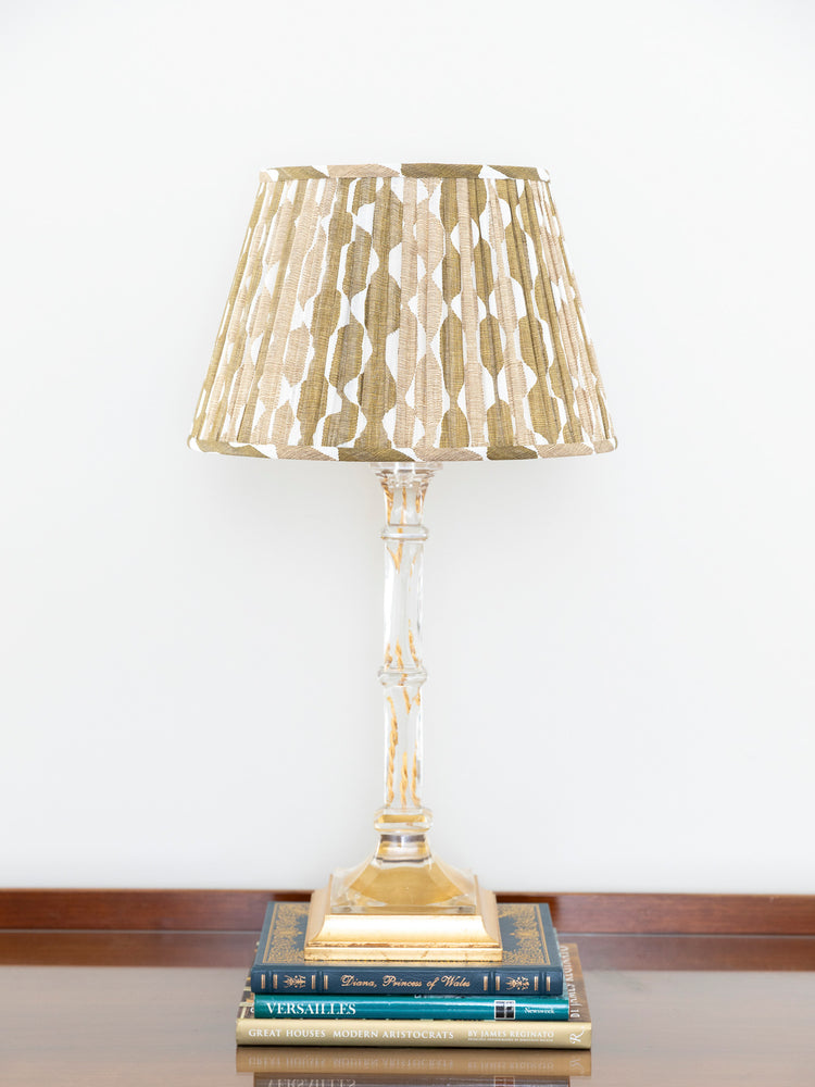A table lamp with a pleated gold and beige shade, a clear glass stem, and a gold base, styled on top of two books against a white wall.